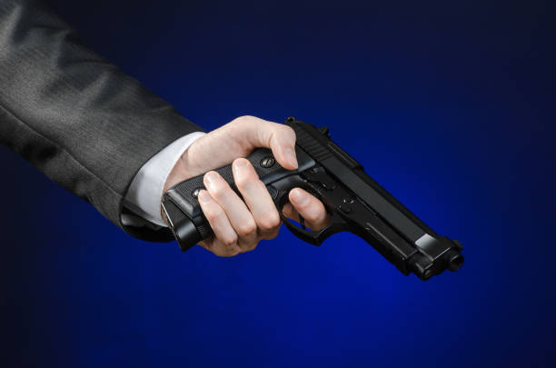armes à feu et la sécurité thème : un homme dans un costume noir, tenant un pistolet sur un fond bleu foncé en studio isolé - gun men spy suit photos et images de collection