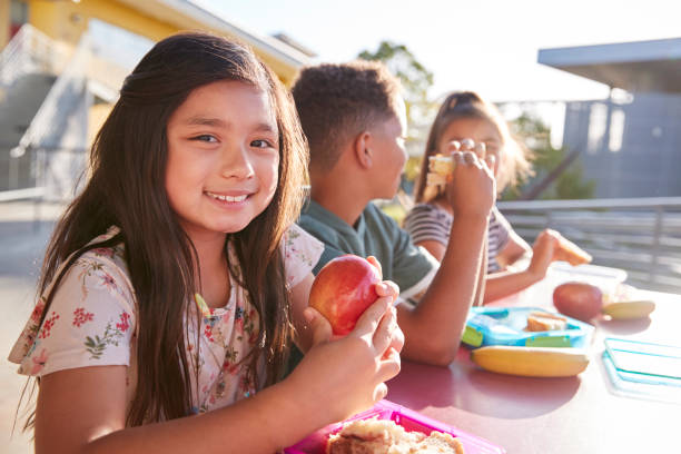girl at elementary school lunch table smiling to camera - child eating imagens e fotografias de stock
