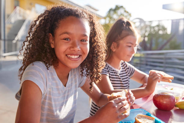 fidanzate a tavola a scuola, una sorridente alla telecamera - child food school children eating foto e immagini stock