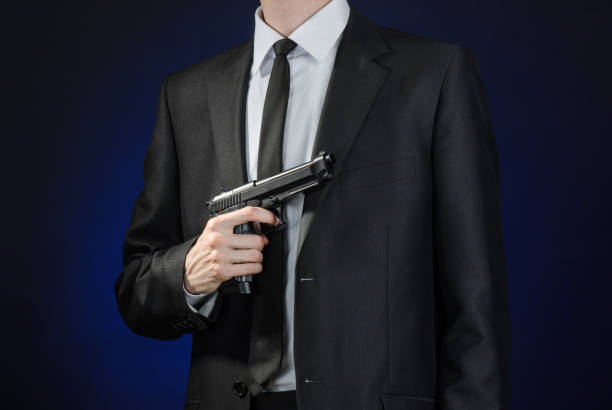 armes à feu et la sécurité thème : un homme dans un costume noir, tenant un pistolet sur un fond bleu foncé en studio isolé - handgun gun blue black photos et images de collection
