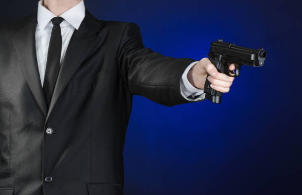 armes à feu et la sécurité thème : un homme dans un costume noir, tenant un pistolet sur un fond bleu foncé en studio isolé - gun men spy suit photos et images de collection