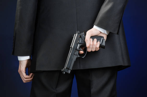 armes à feu et la sécurité thème : un homme dans un costume noir, tenant un pistolet sur un fond bleu foncé en studio isolé - handgun gun blue black photos et images de collection