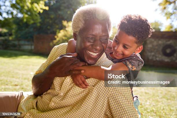 Senior Black Man Sitting On Grass Embraced By His Grandson Stock Photo - Download Image Now