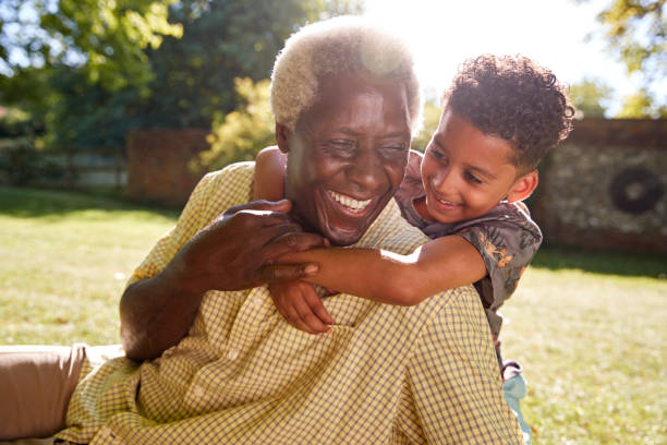 senior black man sitting on grass, embraced by his grandson - environmental portrait imagens e fotografias de stock