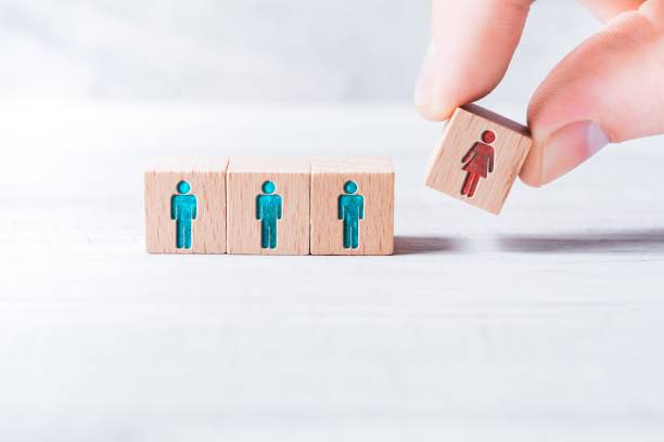 Male Fingers Adding A Block With A Different Colored Female Icon To 3 Blocks With Equal Colored Man Icons On A Table - Gender Equality Concept stock photo