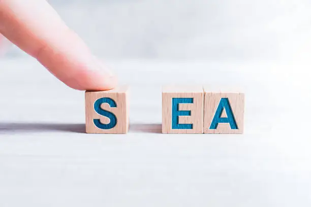 The Word SEA Formed By Wooden Blocks And Arranged By A Male Finger On White Table