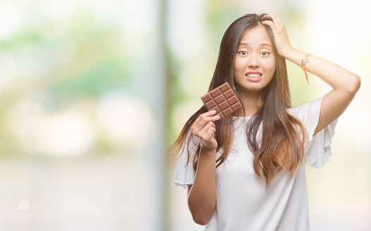 Young asian woman eating chocolate bar over isolated background stressed with hand on head, shocked with shame and surprise face, angry and frustrated. Fear and upset for mistake.