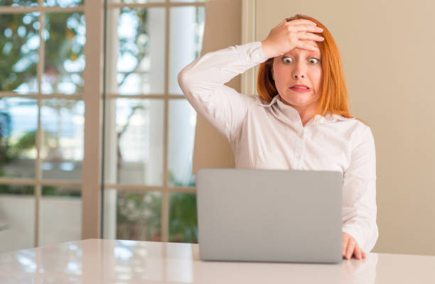 istock Redhead woman using computer laptop at home stressed with hand on head, shocked with shame and surprise face, angry and frustrated. Fear and upset for mistake. 1045397642