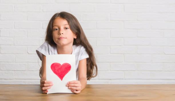 istock Young hispanic kid sitting on the table giving mother day card with a confident expression on smart face thinking serious 1045409340