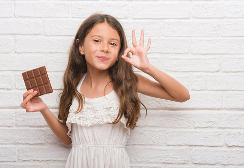 Young hispanic kid over white brick wall eating chocolate bar doing ok sign with fingers, excellent symbol