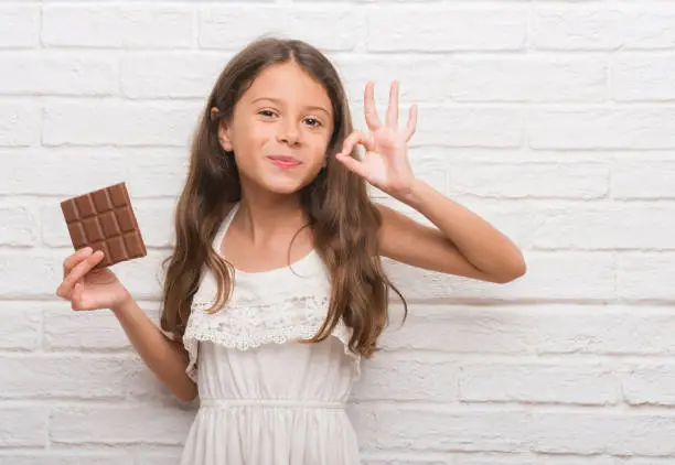 Photo of Young hispanic kid over white brick wall eating chocolate bar doing ok sign with fingers, excellent symbol