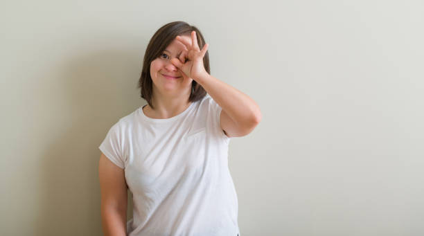 istock Down syndrome woman standing over wall doing ok gesture with hand smiling, eye looking through fingers with happy face. 1045415086