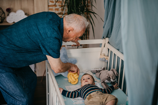 Grandpa playing with his grandson who is laying in a baby cot
