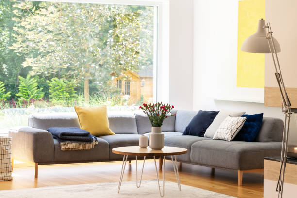 istock Flowers on table next to grey corner sofa with cushions in apartment interior with window. Real photo 1045438222