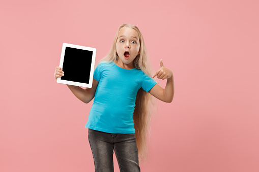 Little funny girl with tablet on pink studio background. She showing something and pointing at screen.