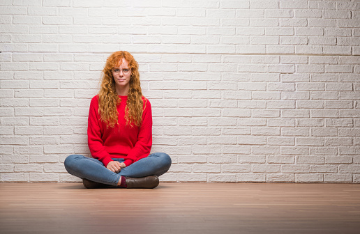 Young redhead woman sitting over brick wall with serious expression on face. Simple and natural looking at the camera.