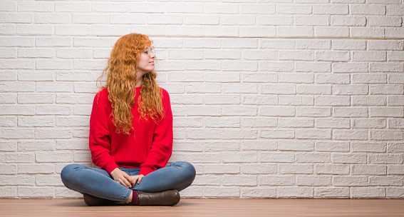 Young redhead woman sitting over brick wall looking to side, relax profile pose with natural face with confident smile.