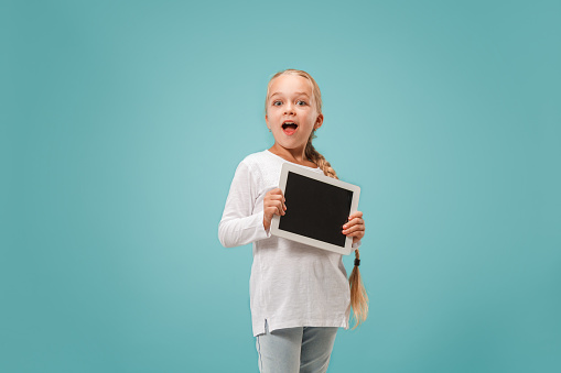 Little funny girl with tablet on blue studio background. She showing something and pointing at screen.