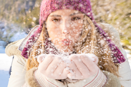 Close-up front view photo of a young woman blowing snow from her hands.