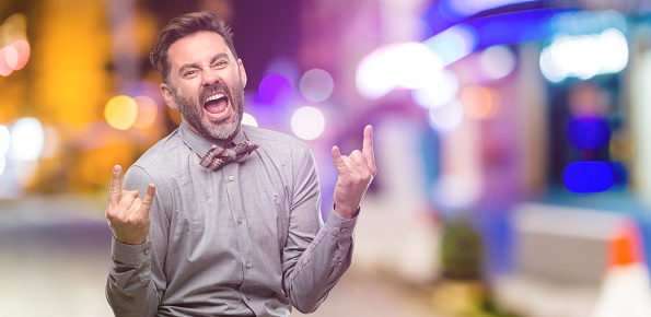 Middle age man, with beard and bow tie making rock symbol with hands, shouting and celebrating at night club