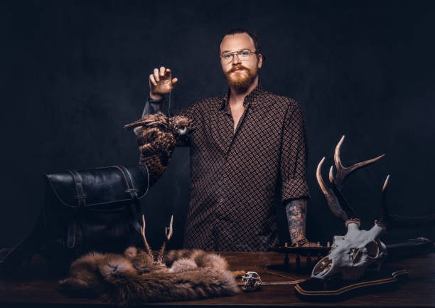 Redhead taxidermist hipster male in sunglasses dressed in a brown shirt, standing near a table with handmade trophy, owl scarecrow, and the fox skin. Isolated on the dark background. Redhead taxidermist hipster male in sunglasses dressed in a brown shirt, standing near a table with handmade trophy, owl scarecrow, and the fox skin. Isolated on a dark background. taxidermy stock pictures, royalty-free photos & images