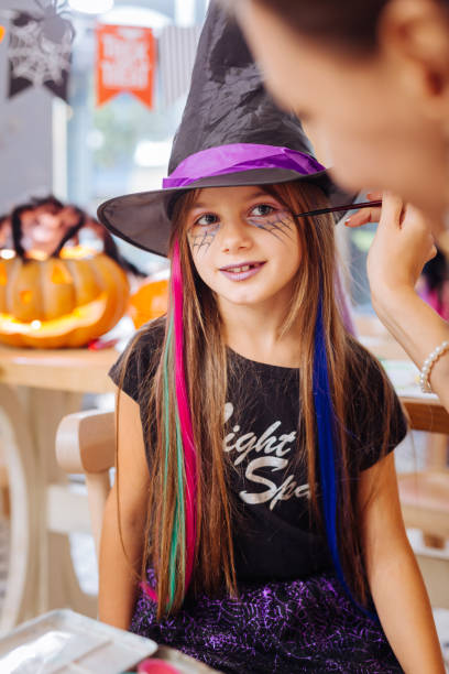 istock Dark-haired girl wearing wizard hat feeling happy getting Halloween makeup 1045504370