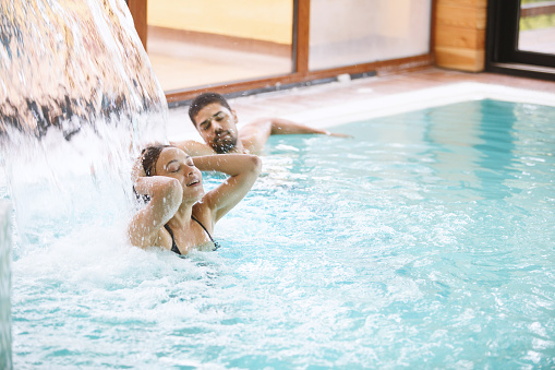 Mixed-race couple are bathing in relaxation swimming pool at the spa. Woman is enjoying under waterfall jet, while her partner is resting at poolside.