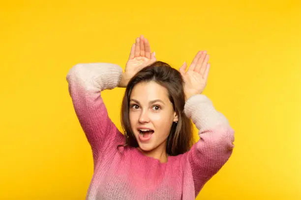 funny amusing playful girl pretending to be a bunny faking ears with her palms. teenage carefree lifestyle concept. young beautiful brown haired woman portrait on yellow background.