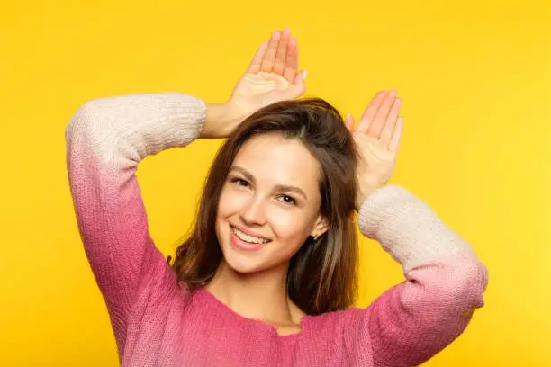 funny amusing playful girl fooling around pretending to be a kitty faking ears with her palms. young beautiful brown haired woman portrait on yellow background.