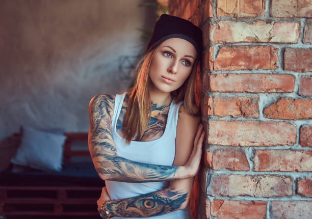 eine tätowierten sexy blondine in einem t-shirt und einem hut posieren gegen eine mauer. - tattoo t shirt brick wall portrait stock-fotos und bilder