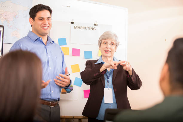 Interpreter signing during business meeting. Multi-ethnic group of business people in team meeting. Interpreter uses sign language to interpret business presentation to a hearing-impaired team member. The team conducts presentation using whiteboard, charts and graphs. sign language stock pictures, royalty-free photos & images