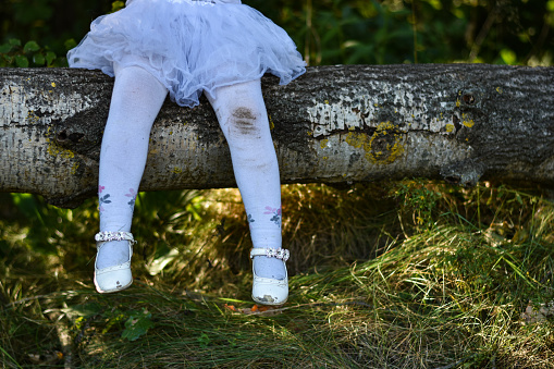 Legs of a little girl in a white dress and dirty pantyhose in the woods, old torn shoes, sitting on tree