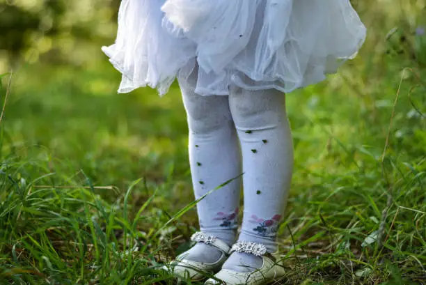 Legs of a little girl in a white dress and pantyhose on the grass in the forest are stained in the ground, grass and thorns, old torn shoes