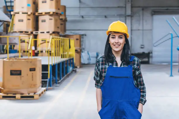 Portrait of a young female trainee wears blue coverall and leans, look at camera posing while standing, smiling, working in metal industry large shipping manufacturing facility warehouse at metal cable factory with copy space area. XXXL size