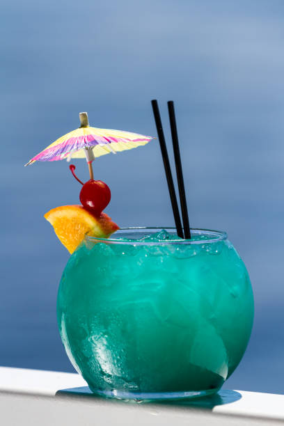 blue cocktail served in a fish bowl served on a railing with a natural water background as a travel concept