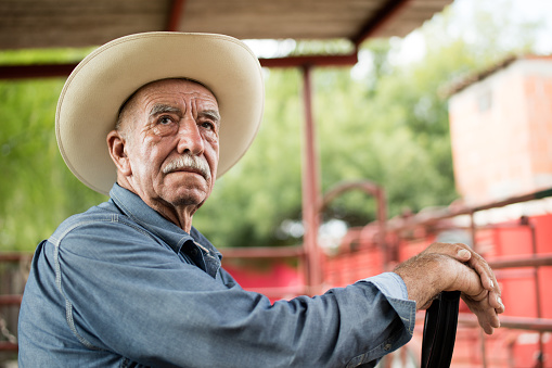 A senior mexican man wearing a cowboy hat, leaning his hands and looking away.