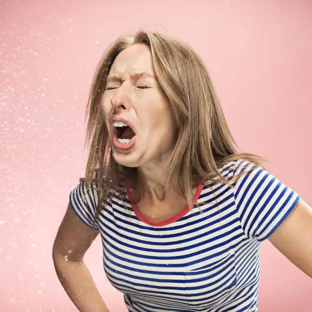 Young funny woman sneezing with spray and small drops, studio portrait on pink background. Comic, caricature, humor. illness, infection, ache. Health concept