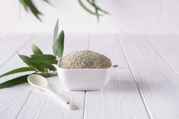 istock Ceramic bowl with green clay powder and fresh eucalyptus leaves on white background. Concept of face and body care. 1045843232