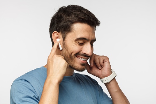 Young man wearing wireless earbuds and blue t shirt, listening to his favorite musical album online, touching one earphone to control application