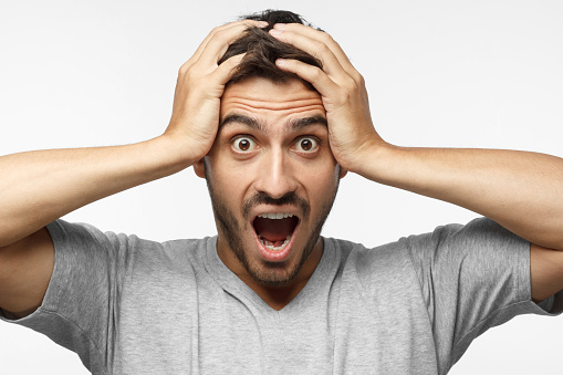 Close up shot of shocked young man with round eyes, open mouth and hands pressed to top of head, showing astonishment and shock, isolated on gray background