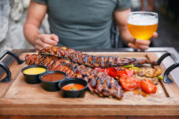 Barbecue Meat With Vegetables And Beer In Restaurant. Man Eating Grill Pork Ribs Closeup. High Resolution