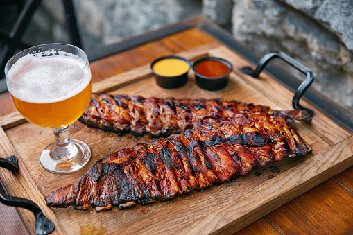 Barbecue Ribs And Beer On Table In Restaurant Closeup. Spareribs With Grilled Vegetables And Sauce On Wooden Tray. High Resolution