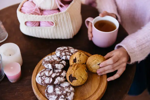 Cup of black tea and plate of fresh homemade cookies on wooden table with woolen coils of thread. Woman hands holding hot aromatic tea. Morning breakfast, warm and cozy