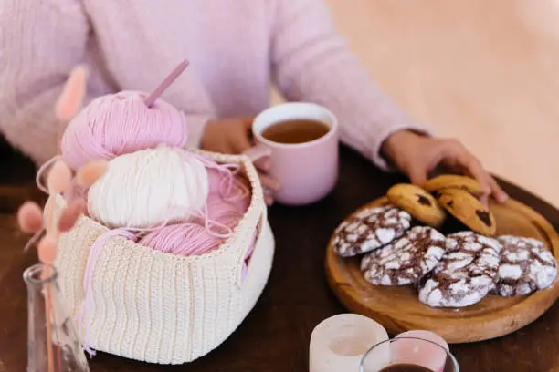 Cup of black tea and plate of fresh homemade cookies on wooden table with woolen coils of thread. Woman hands holding hot aromatic tea. Morning breakfast, warm and cozy