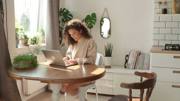 Charming young woman typing on laptop computer in a kitchen. stock photo