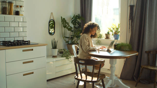 Charming young woman typing on laptop computer in a kitchen. stock photo