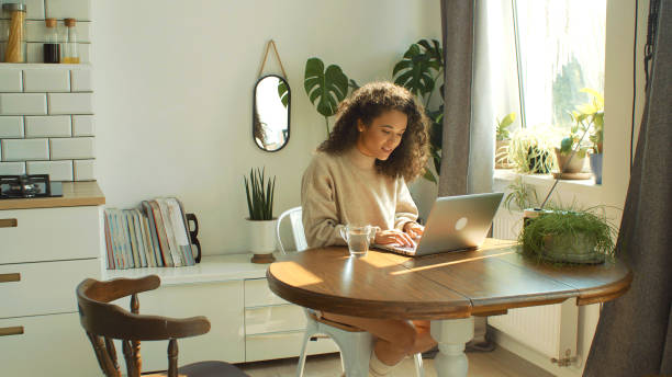 encantadora mujer joven escribiendo en la computadora portátil en una cocina. - women computer home interior brown hair fotografías e imágenes de stock