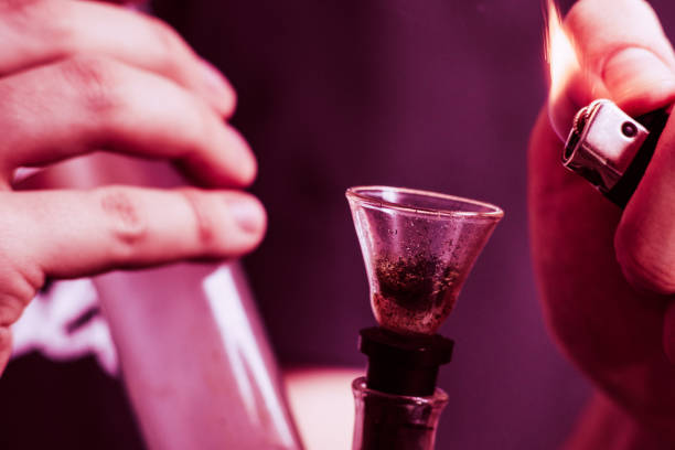 istock Bong lighter for A man smokes cannabis weed, in his hands. Smoke on a black background. Concepts of medical marijuana use and legalization of the cannabis. On a black background Violet tinting 1045971214