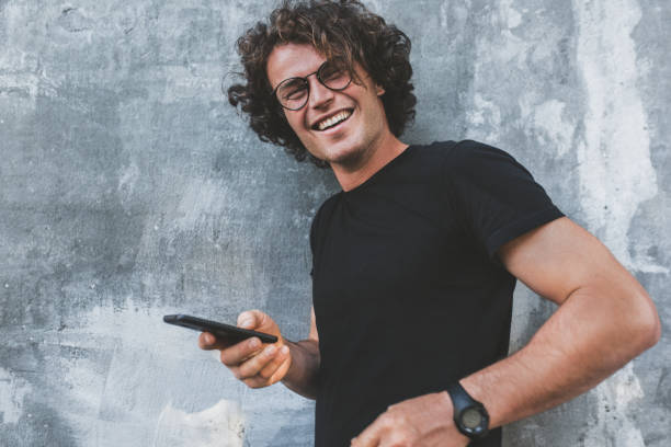 istock Handsome happy smiling man standing outdoors texting on mobile phone. Young male with curly hair wears spectacles resting outside in the city browsing on his cell phone on concrete gray background. 1045975130