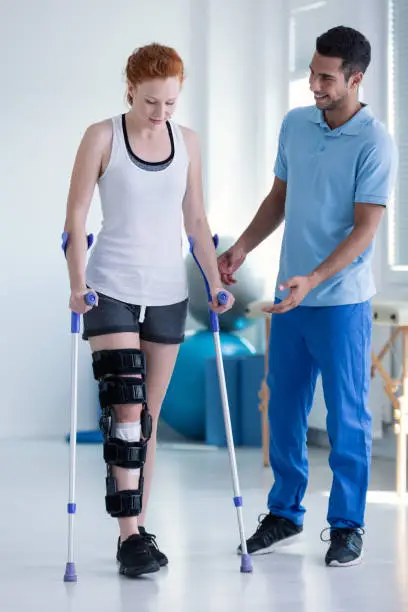 Photo of Physiotherapist helping woman with stiffener on the leg walking with crutches